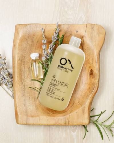 Organethic Pure Care Wellness Energising Shampoo in a clear bottle on a wooden tray with lavender and rosemary